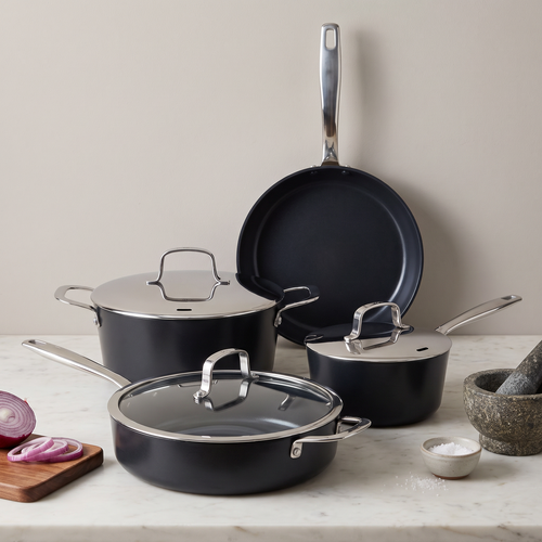 A cookware set arranged on a light stone countertop against a warm beige wall, including dark nonstick pots and pans with silver metal handles and lids, plus a cutting board with sliced red onion, a small bowl, and a stone mortar in the background.