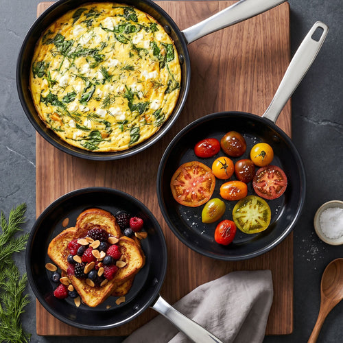 Three black nonstick pans with silver handles arranged on a wooden board, containing cooked foods: a large omelet with green herbs, sliced tomatoes, and fruit, toast topped with berries and nuts, and mixed cherry tomatoes. A small bowl of salt, a wooden spoon, and a folded gray cloth are visible on a dark surface.